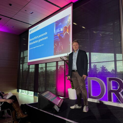 Ein Mann steht auf einer Bühne und hält einen Vortrag, im Hintergrund eine Leinwand