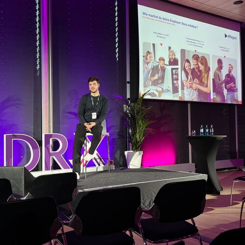 Ein Mann sitzt auf einem Hocker auf einer Bühne und spricht zu einem Publikum, während auf einer Leinwand