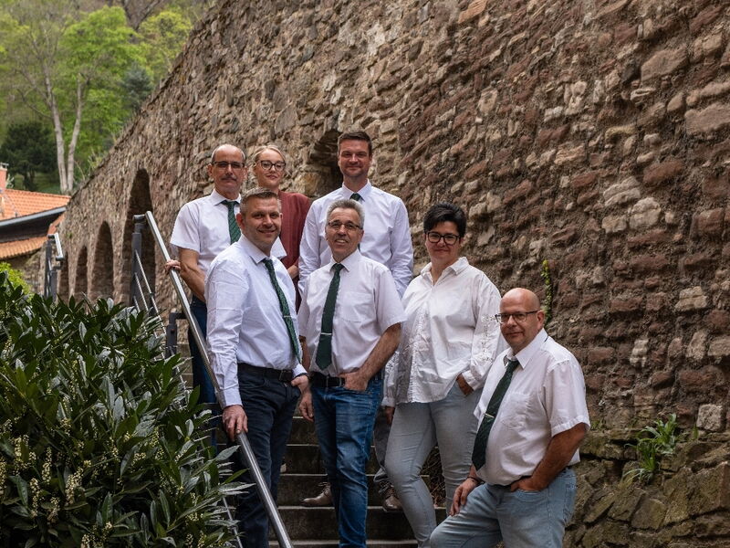 Sieben Personen in weißen Hemden und Krawatten stehen auf einer Steintreppe vor einer alten Steinmauer