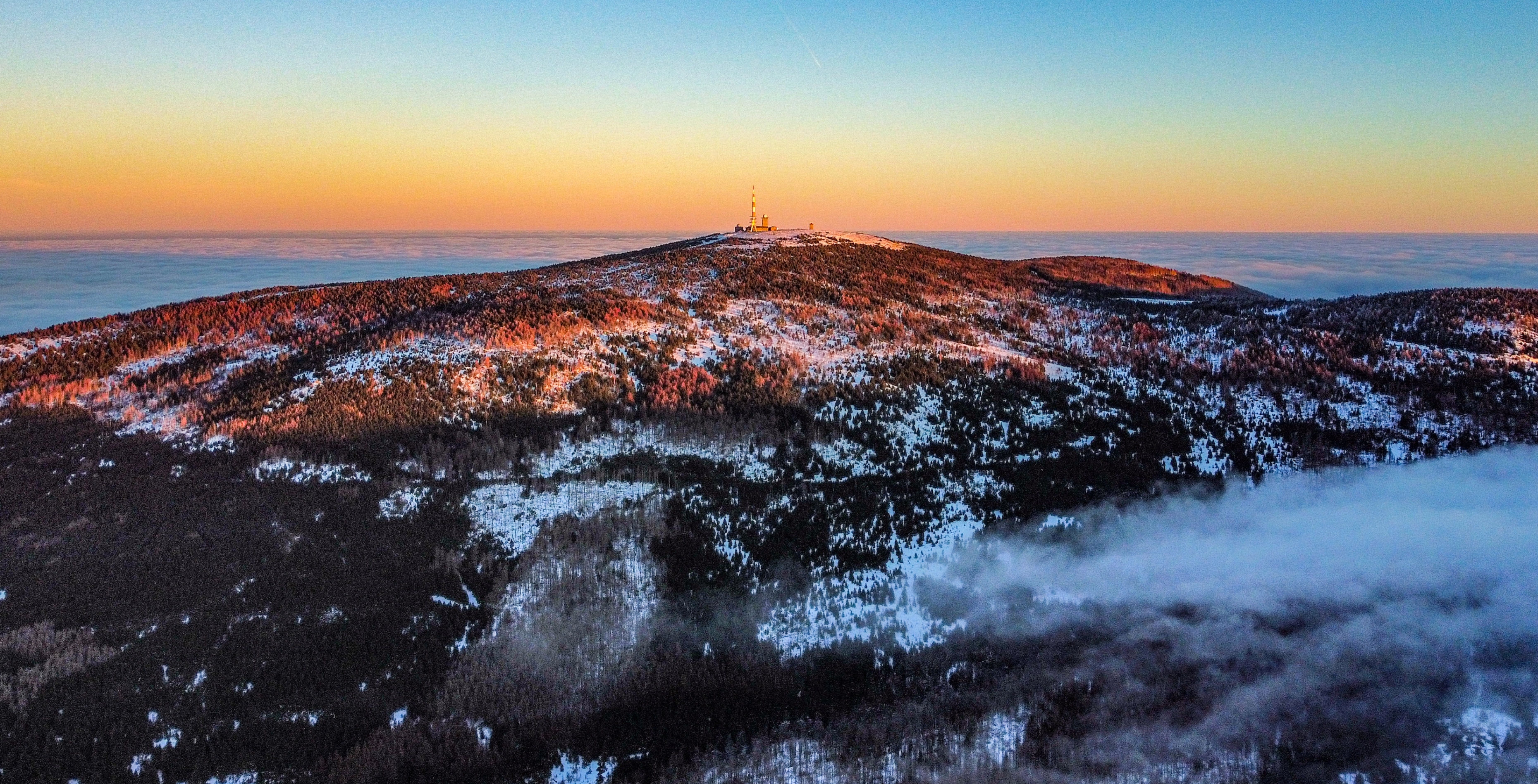 Ein Berg mit einem Sendeturm auf dem Gipfel, bedeckt mit Schnee und Bäumen, im warmen Licht des